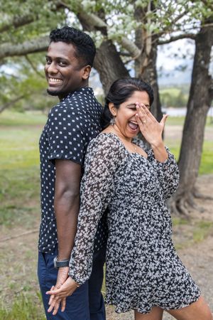 A playful couple laughs together during an outdoor photoshoot with trees in the background.
