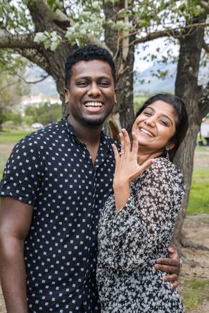 A joyful couple poses outdoors showing off an engagement ring amid trees and natural scenery.