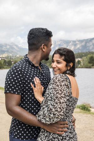 A couple embraces by a mountain lake, with the woman showing off her ring against a scenic backdrop.