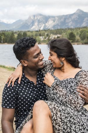 A romantic couple laughs together while sitting lakeside with mountains in the distance.