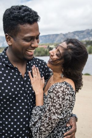 A couple shares a candid laughing moment against a mountain lake backdrop.