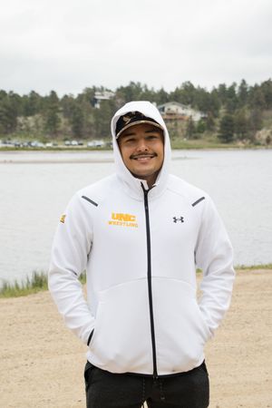 Person in a white Under Armour hoodie smiling by a lakeside with mountains in the background.