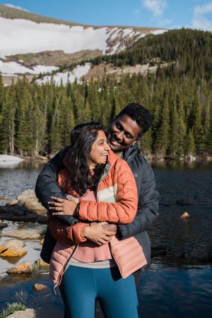 Couple embracing by a scenic mountain lake with snow-capped peaks and evergreen forest.
