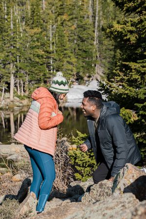Two figures in winter coats explore rocky outcrops by an alpine lake in a forest setting.