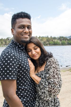 A couple shares an intimate moment while posing together near a waterfront with mountains in the background.