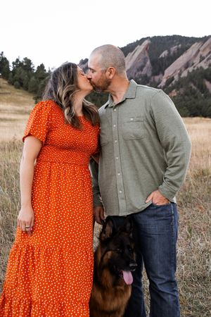 A couple in orange dress and olive shirt pose together with their black dog against scenic mountain backdrop.