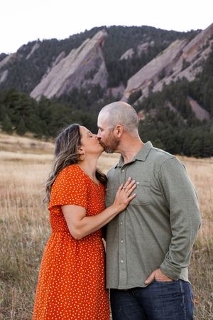 A couple shares a romantic kiss in a field with mountains and Flatirons rock formations in the background at sunset.
