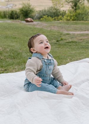 Happy baby in Levi's denim overalls sitting on white blanket in green outdoor setting.