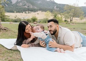 Family with baby lying on white blanket in scenic mountain meadow during outdoor photo session.