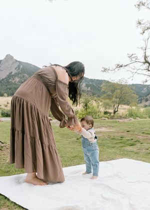 A mother in a brown dress helps her toddler walk on a white blanket with mountains in the background.