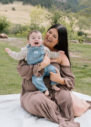 A laughing mother in a brown dress sits on a blanket holding her joyful baby in denim overalls outdoors.