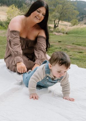 A smiling mother watches her baby crawl on a white blanket during an outdoor photo session in a green meadow.