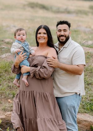 A smiling family of three poses outdoors, with a woman in a brown dress and a man holding a baby in denim.