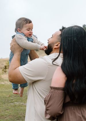 A father lifts his smiling baby outdoors while a woman with long dark hair stands nearby in a grassy field.