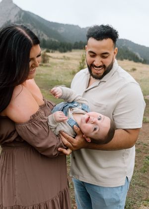A joyful family with a laughing baby outdoors near mountains, parents smiling and playing with their infant child.