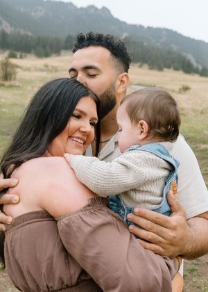 A couple smiles while holding their baby outdoors in a green mountain meadow during a family photo session.