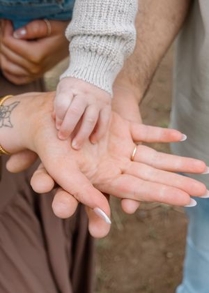 A baby's tiny hand resting on adult palms, one wearing a gold wedding band and a tattoo visible on the wrist.