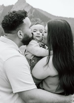 Black and white photo of parents kissing their baby on the cheeks with mountains in the background.