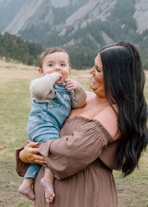 A mother in a brown off-shoulder dress holds her baby boy in denim overalls with Rocky Mountains in the background.