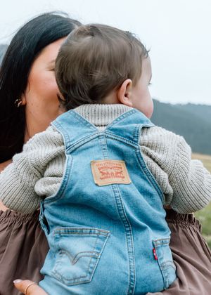 Mother kissing baby wearing Levi's denim overalls and beige knit sweater outdoors.