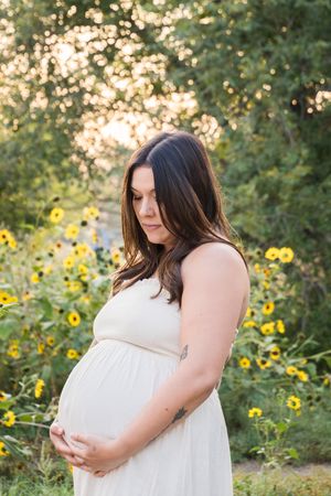 Expecting parent stands in a flowing white dress in a garden with sunflowers during golden hour.