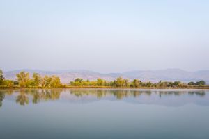 Calm lake reflects tree line and distant mountains under a hazy sky during golden hour.
