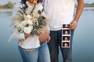Couple holding ultrasound photos shares pregnancy announcement at scenic lakeside location in casual attire.
