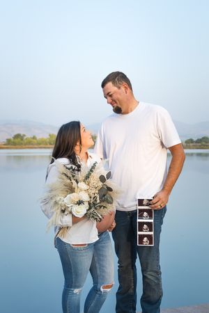 Couple poses lovingly at waterfront location sharing pregnancy ultrasound photos and embracing with natural scenery behind them.