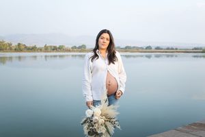 A person in a white sweater holds dried flowers while standing by a serene lake on an overcast day.