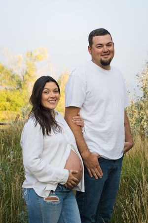 Expecting couple in coordinating white tops and jeans pose together in tall grass during outdoor maternity photo session.
