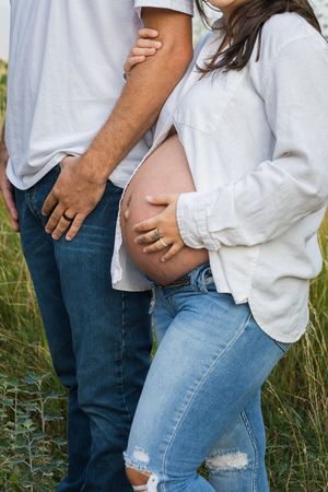 Couple in casual denim and white shirts embracing while showing pregnant belly in outdoor field setting.