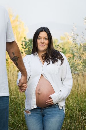 A pregnant person in a white button-down shirt and jeans poses in a sunlit field of tall grass.