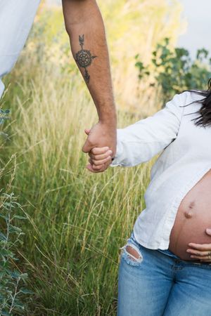 A pregnant person in blue jeans and white top poses against a natural outdoor backdrop with tall grasses.
