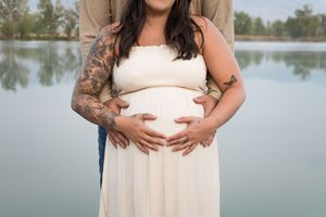 Maternity photo shoot by a lake showing an expecting mother in a flowing cream dress cradling her baby bump.