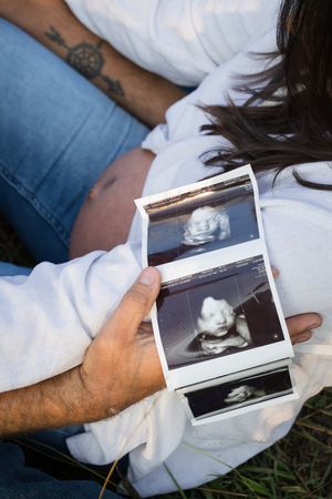 Someone wearing jeans and a white sweater holds ultrasound sonogram photos while sitting on grass.