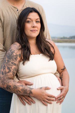 Expectant couple poses outdoors near water, with person in white dress showing baby bump and floral sleeve tattoo.