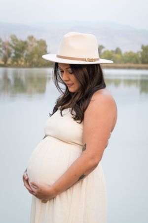 A pregnant woman in a cream dress and white hat poses by a lake during a maternity photoshoot.