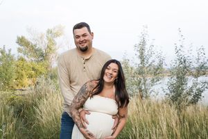 Expectant couple in casual attire embraces in a sunlit field of tall grass during golden hour maternity photo session.