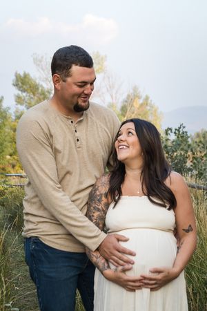 A loving couple in neutral colors share joyful moments during their maternity photo session by a lake.