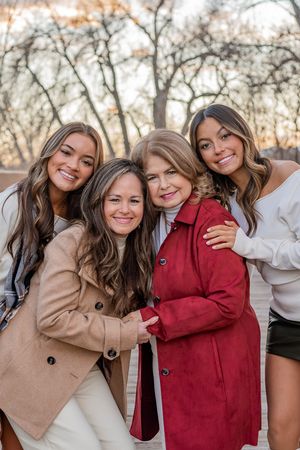 A group of women in coats sharing a warm embrace outdoors during golden hour with bare trees in the background.