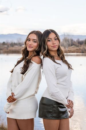 Two people in white sweaters pose together near a lake with mountains in the background.