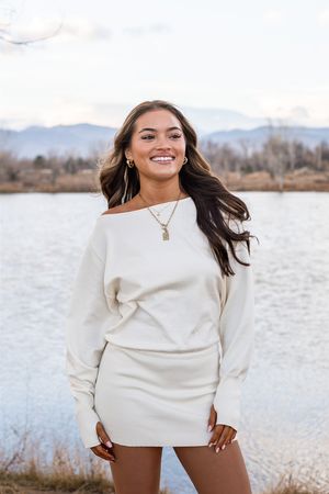 A woman in a cozy cream-colored sweater dress poses outdoors by a lake with mountains in the background.
