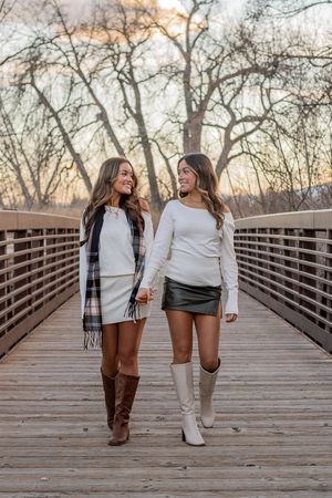 Two friends walking together on a wooden bridge during sunset in autumn wearing cozy sweaters and boots.