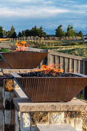 Two square metal fire pits with orange flames sit atop stone bases on an outdoor patio with wooden railings.