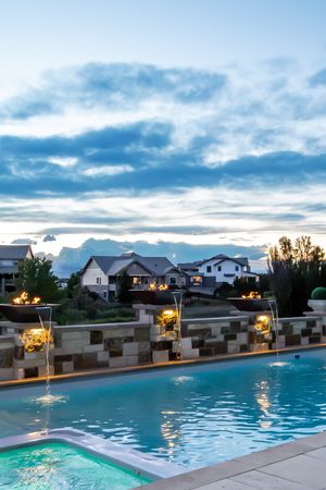 Elegant backyard pool with water features at dusk, fire bowls, and suburban homes under dramatic cloudy sky.