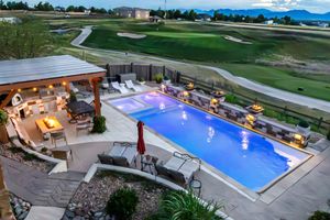 Aerial view of a luxury backyard with a glowing pool, outdoor kitchen, pergola, and golf course views at dusk.