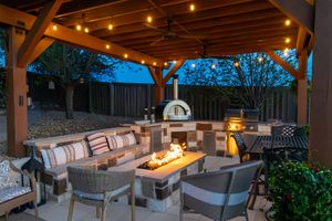 Cozy covered outdoor patio with string lights, fire pit table, seating, and a wood-fired pizza oven at dusk.