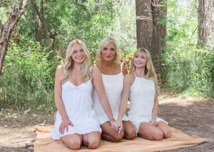 Three women in white dresses pose together while sitting on a wooden platform in a forest setting.