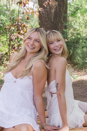 Two women in white dresses share joyful laughter while sitting together outdoors in a wooded setting.