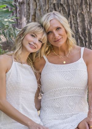 Two people pose together outdoors wearing white lace dresses against a tree trunk background.
